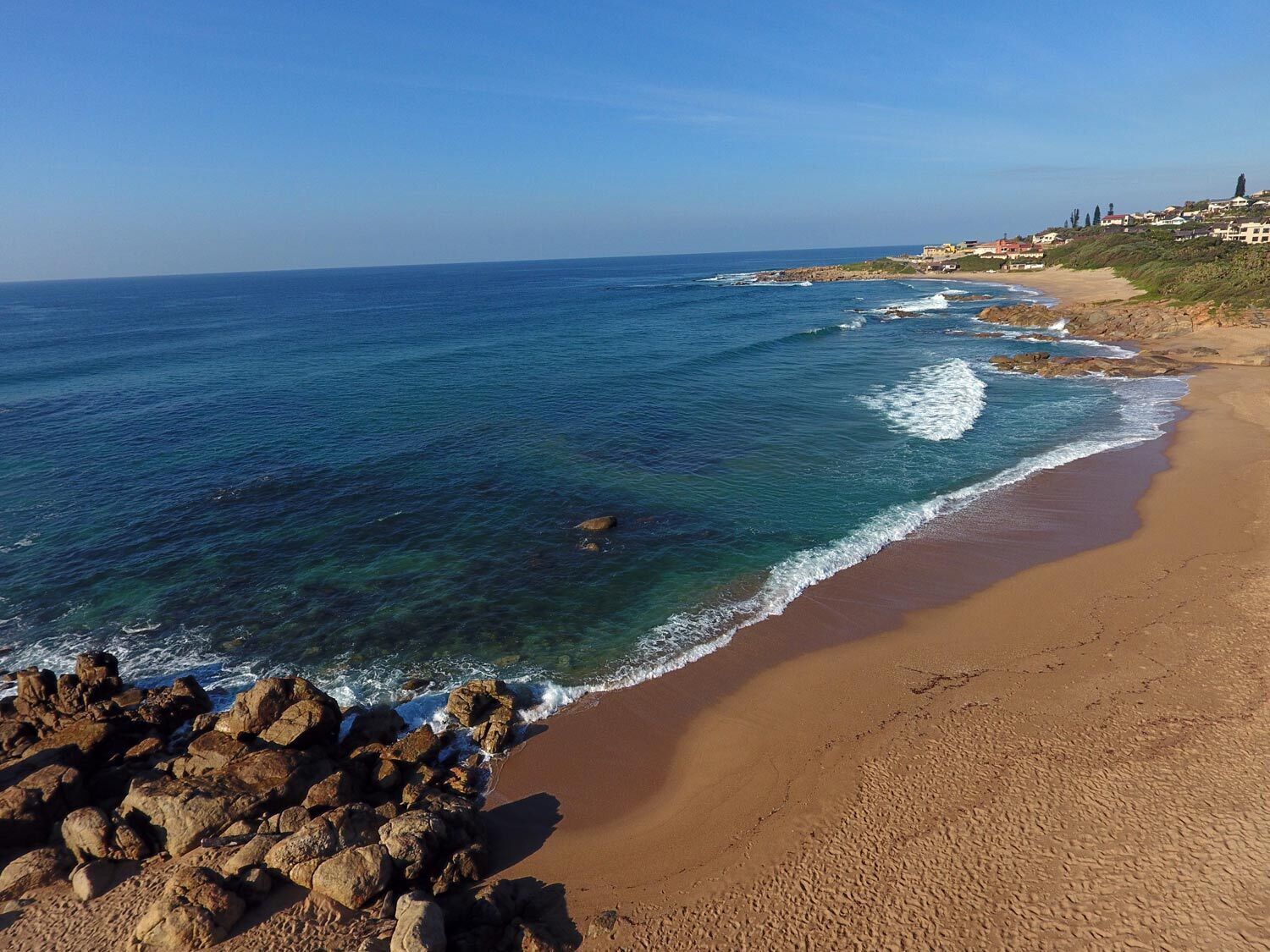 Une plage côtière aux eaux bleues et limpides, avec un rivage sablonneux et des affleurements rocheux.
