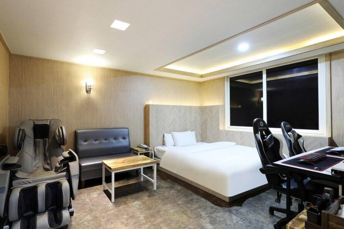 A hotel room with a bed, a desk with a chair, a massage chair, and a large window.