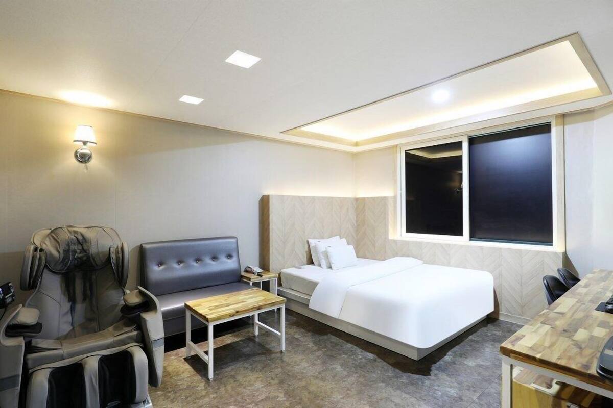 A hotel room with a bed, a sofa, a massage chair, a desk, and a window.