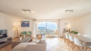 A modern living room with a large TV, sofa, dining table, and chairs. There is a balcony with a view of the sea. - Villa la Cagne (Saint-Raphaël)