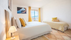 A bedroom with a large bed, a smaller bed, a nightstand with a lamp, a window with curtains, and a painting on the wall. - Villa la Cagne (Saint-Raphaël)