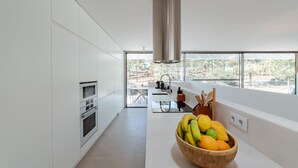 A modern kitchen with white cabinetry, a stainless steel range hood, a wooden bowl of fruit on the counter, and a view of the outdoors through large windows.