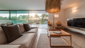 A modern living room with a sofa, coffee table, and TV. There is a large window with a view of trees and a balcony with chairs.