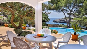 A covered outdoor area with a pool, white chairs, and a table with fruit.
