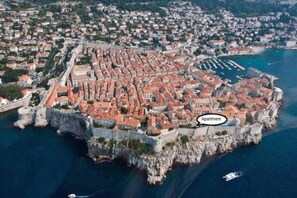 An aerial view of a coastal town with a prominent fortress-like structure surrounded by red-roofed buildings, a marina with boats, and a label indicating an "Apartment."