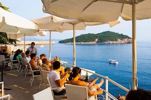 An outdoor dining area with tables and chairs under umbrellas, overlooking a body of water and a hilly landscape.