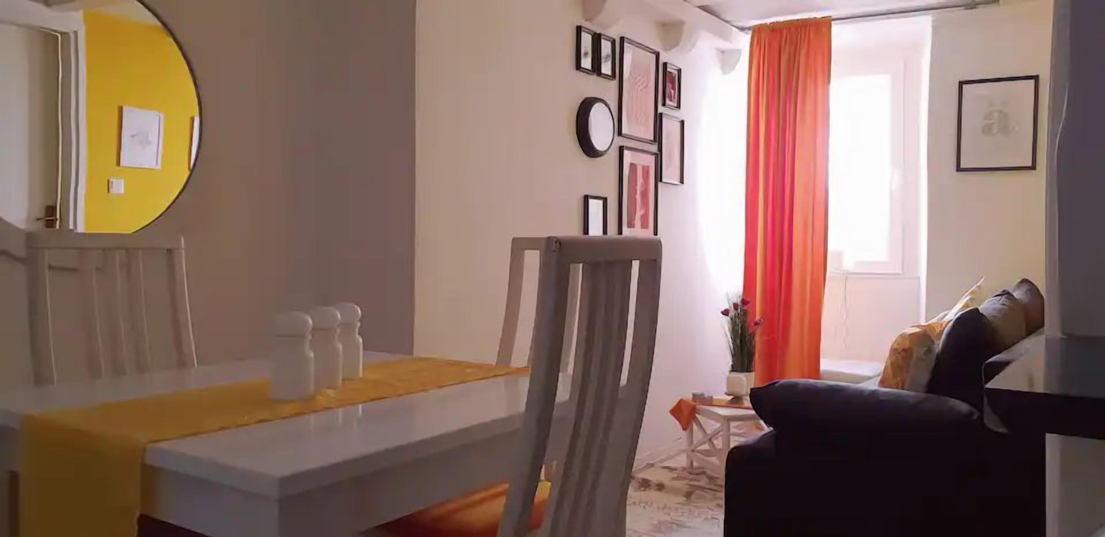 A dining area with a table, chairs, and a yellow table runner. There is a sofa and a window with curtains in the background.