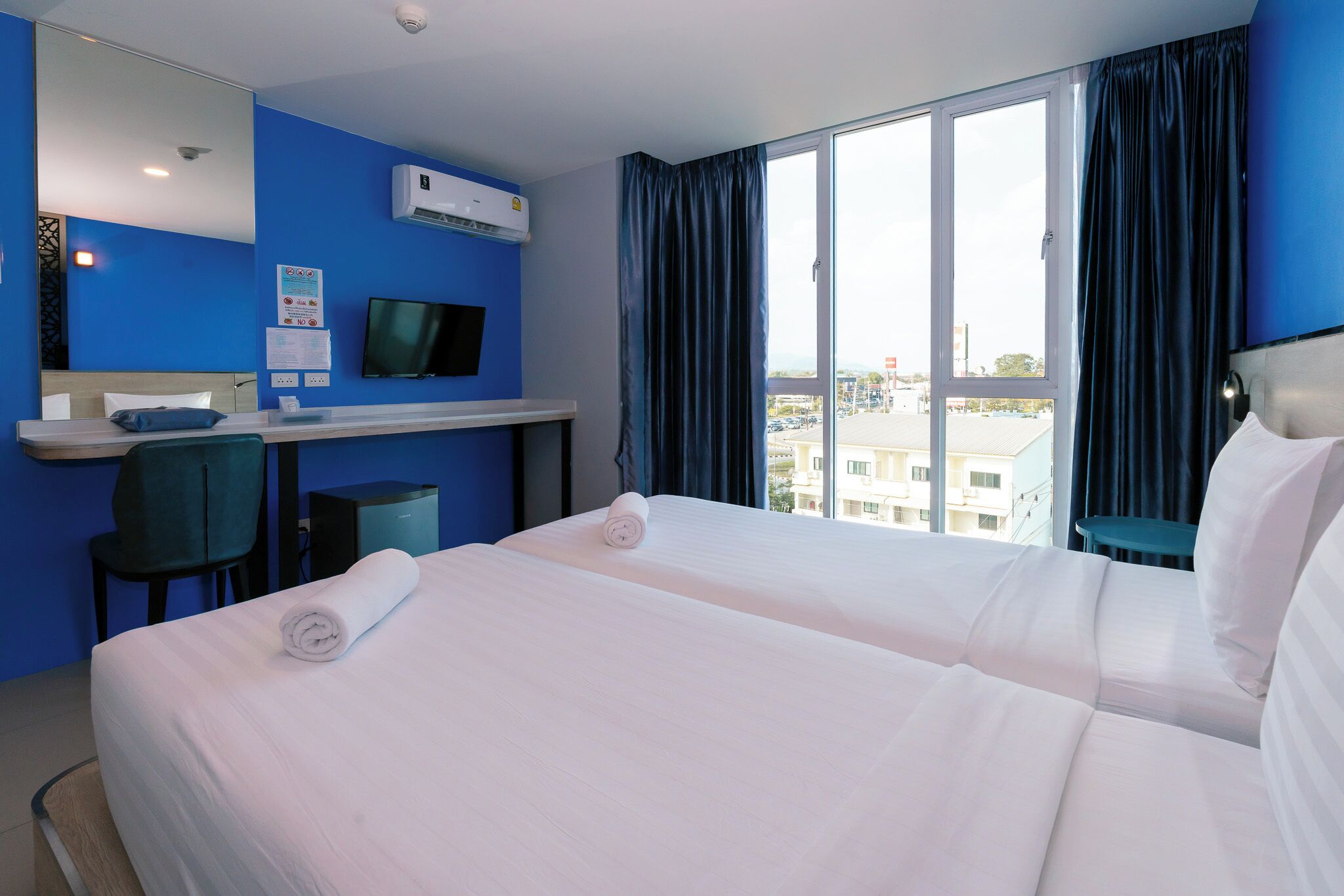 A hotel room with two beds, a desk, a chair, a TV, and a large window with curtains.