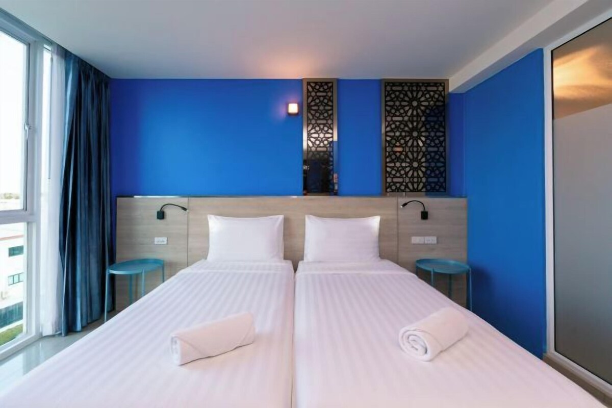 Habitación de hotel con dos camas, un ventanal grande, pared azul y un panel decorativo.