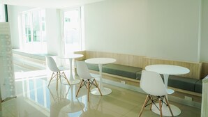 Un comedor moderno con mesas y sillas blancas, un banco y ventanas amplias que dejan entrar luz natural.