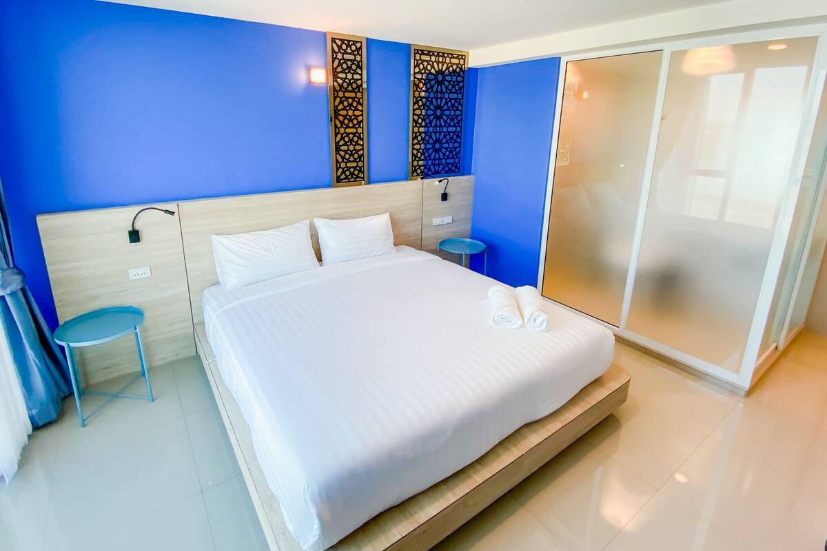 Una habitación de hotel con una cama grande, una mesita de noche, un taburete pequeño, un armario y un panel decorativo en la pared.
