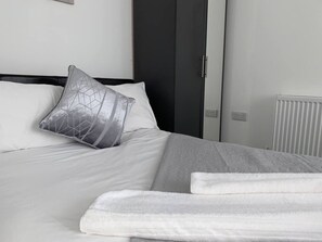Een net opgemaakt bed met witte lakens en een kussen met een geometrisch patroon, een witte radiator en een donkere deur in een moderne kamer.