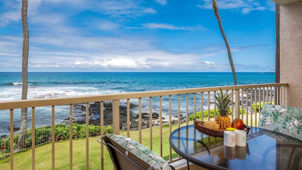 A balcony with a view of the ocean, a table with fruits and a basket, and a chair.