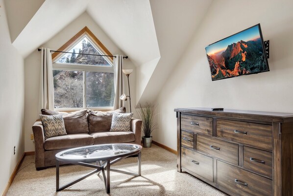 Ein gemütlicher Raum mit schrägen Wänden, einem großen Fenster mit Blick auf Bäume, einem braunen Sofa mit gemusterten Kissen, einem Glastisch, einer Holzkommode und einem gerahmten Bild an der Wand.