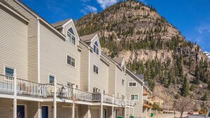 A multi-story building with a balcony on the ground floor, a mountain backdrop, and a parked vehicle in the foreground. - Near Hot Springs + Downtown Ouray | Deck with Views (Ouray)