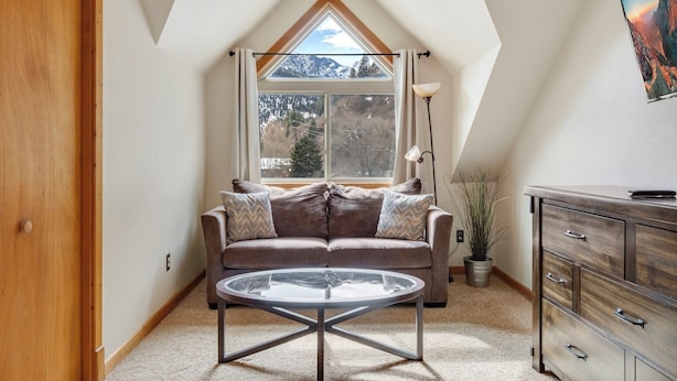 A cozy loft bedroom with a sloped ceiling, a large window offering a mountain view, a brown leather sofa, a glass coffee table, and a wooden dresser. - Near Hot Springs + Downtown Ouray | Deck with Views (Ouray)