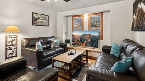 A modern living room with leather sofas, a wooden coffee table, and a large flat-screen TV displaying a mountain view. - Near Hot Springs + Downtown Ouray | Deck with Views (Ouray)