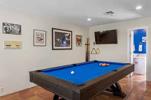 Uma mesa de bilhar com superfície de feltro azul, uma televisão fixada na parede e objetos de coleção emoldurados na parede.