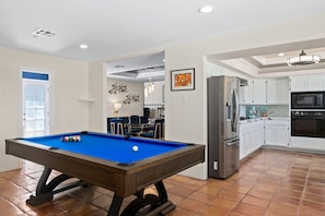 Uma mesa de bilhar em uma sala com cozinha, armários brancos e área de jantar com cadeiras.