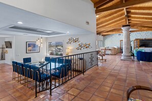Uma ampla área de jantar com mesa de vidro e cadeiras azuis, um pilar branco grande e piso ladrilhado.