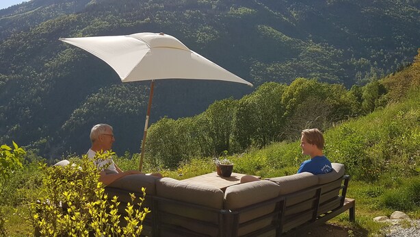 Deux personnes sont assises à une table à l’extérieur, sous un parasol blanc, et elles ont une vue sur un paysage montagneux.