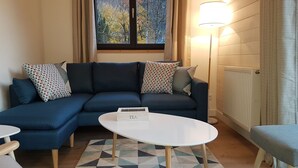 Un salon moderne avec un canapé d’angle bleu, des coussins à motifs, une table basse blanche et une vue sur les arbres par la fenêtre.