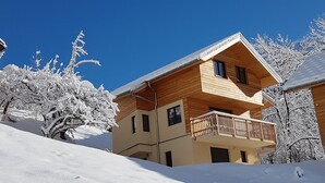 Un chalet en bois à deux étages, doté d’un balcon et entouré d’arbres couverts de neige.