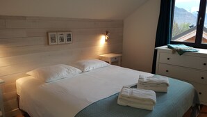 Une chambre à coucher avec une tête de lit en bois, un lit recouvert de draps blancs, une table de chevet avec une lampe, une commode et une fenêtre donnant sur la montagne.