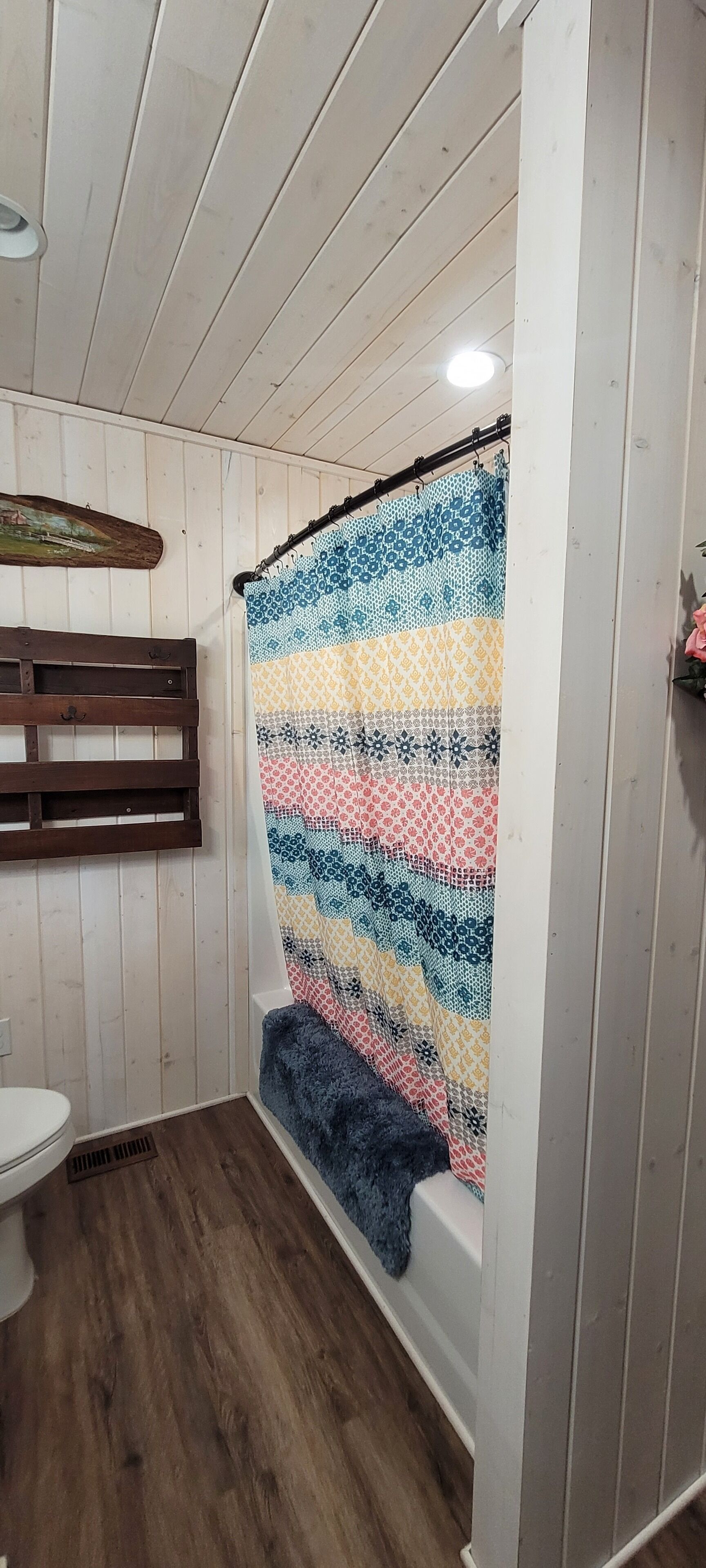 A bathroom with wooden panel walls, a white toilet, a patterned shower curtain, and a wooden bench.
