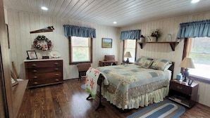 Une chambre avec un plancher en bois, un lit recouvert d’une couvre-lit à motifs floraux, une commode, une table de chevet avec une lampe et une fenêtre agrémentée d’un lambrequin bleu.