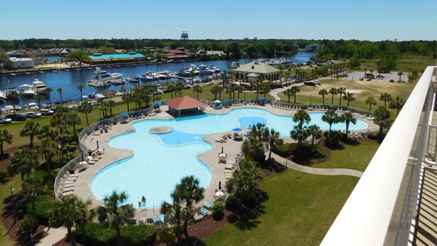 A resort with a large, winding pool surrounded by palm trees and lounge chairs, overlooking a marina with boats and a water tower in the distance. - Yacht Club Villas #1-1005 Beautiful Waterfront Villa in Barefoot Resort! (North Myrtle Beach)