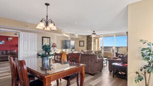A spacious living area with a dining table, chairs, a sofa, and a balcony with a view. - Yacht Club Villas #1-1005 Beautiful Waterfront Villa in Barefoot Resort! (North Myrtle Beach)