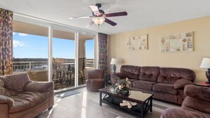 A spacious living room with a brown sofa, a smaller armchair, and a coffee table. There is a ceiling fan and a view of the outdoors through large windows. - Yacht Club Villas #1-1005 Beautiful Waterfront Villa in Barefoot Resort! (North Myrtle Beach)