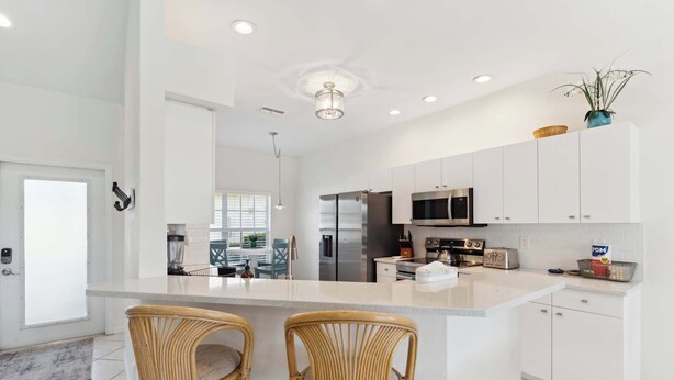 A modern kitchen with white cabinets, stainless steel appliances, and a central island with two wicker chairs. - Poolside Bliss and a Family-Friendly Oasis (Naples)