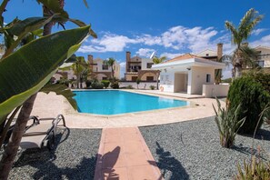 A swimming pool with a clear blue water, surrounded by a paved area with stepping stones leading to a white building with a tiled roof.