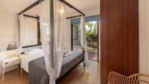 A bedroom with a four-poster bed, a bedside table with a lamp, and a view of the outdoors.