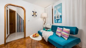 Een woonkamer met een turquoise bank, een klein wit tafeltje met fruit, een lamp en een schilderij van een zeilboot.