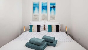 Un lit bien fait, recouvert d’une couette blanche et turquoise, agrémenté de serviettes turquoise, et un mur orné de trois tableaux encadrés représentant une scène de plage.