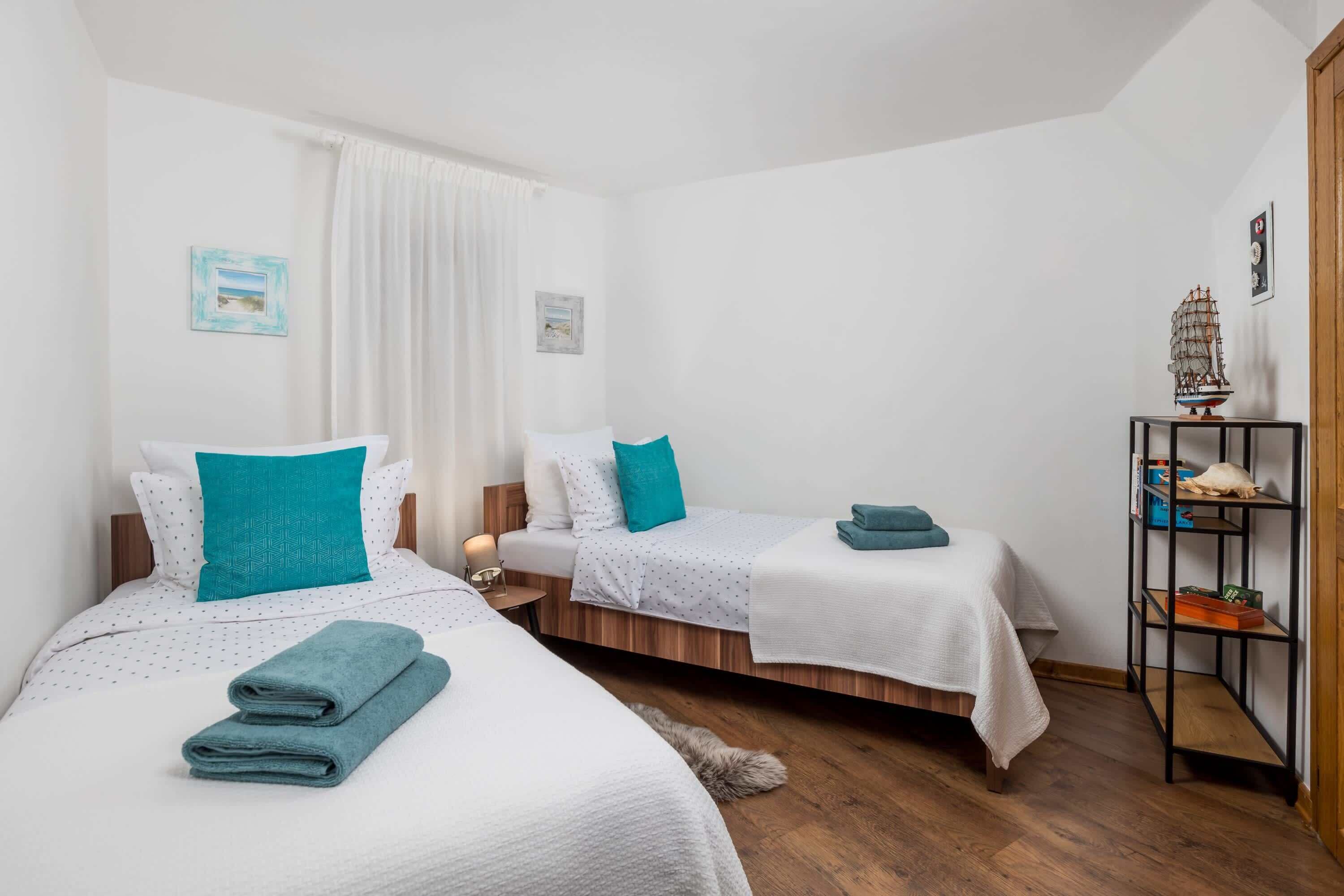 A hotel room with two beds, a wooden floor, a nightstand with a lamp, and a shelf with decorative items.