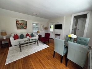 Ein Wohnzimmer mit einem weißen Sofa, roten und türkisfarbenen Kissen, einem Glastisch und einem roten Sessel. Ein Holzboden, ein weißer Teppich und eine Tür zu einem anderen Raum sind ebenfalls zu sehen.
