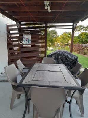 Un'area pranzo all'aperto coperta con tavolo e sedie in legno, un barbecue e un pannello decorativo con un poster.