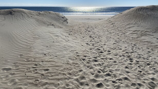 A sandy beach with footprints leading to the water, a clear sky, and the ocean in the background. - Great location! Fully fenced in yard and expansive deck. Come and relax! (Oak Island)