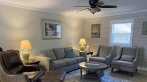 A living room with a grey sofa, two armchairs, a coffee table, and a rug. There are two lamps on side tables, a ceiling fan, and artwork on the walls. - Great location! Fully fenced in yard and expansive deck. Come and relax! (Oak Island)