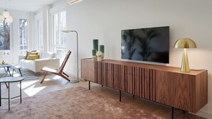 Una sala de estar moderna con un televisor de pantalla plana grande montado en un mueble de madera, una lámpara de pie y un sofá con cojines.