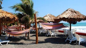 Ein Strand mit Reetdachschirmen, Hängematten und weißen Liegestühlen.