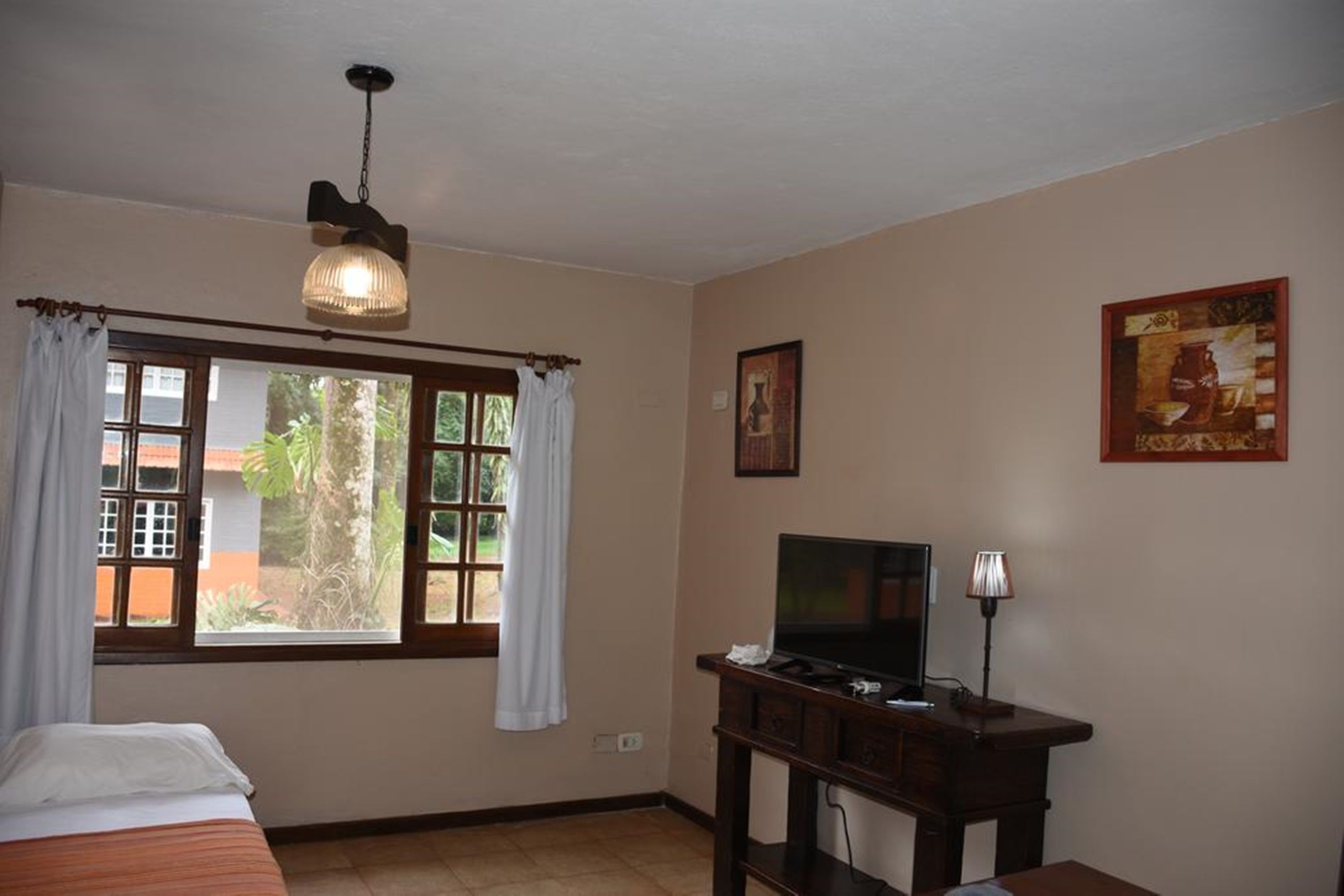 Une chambre d’hôtel avec un lit, un téléviseur posé sur un meuble en bois, une fenêtre avec des rideaux et un tableau accroché au mur.