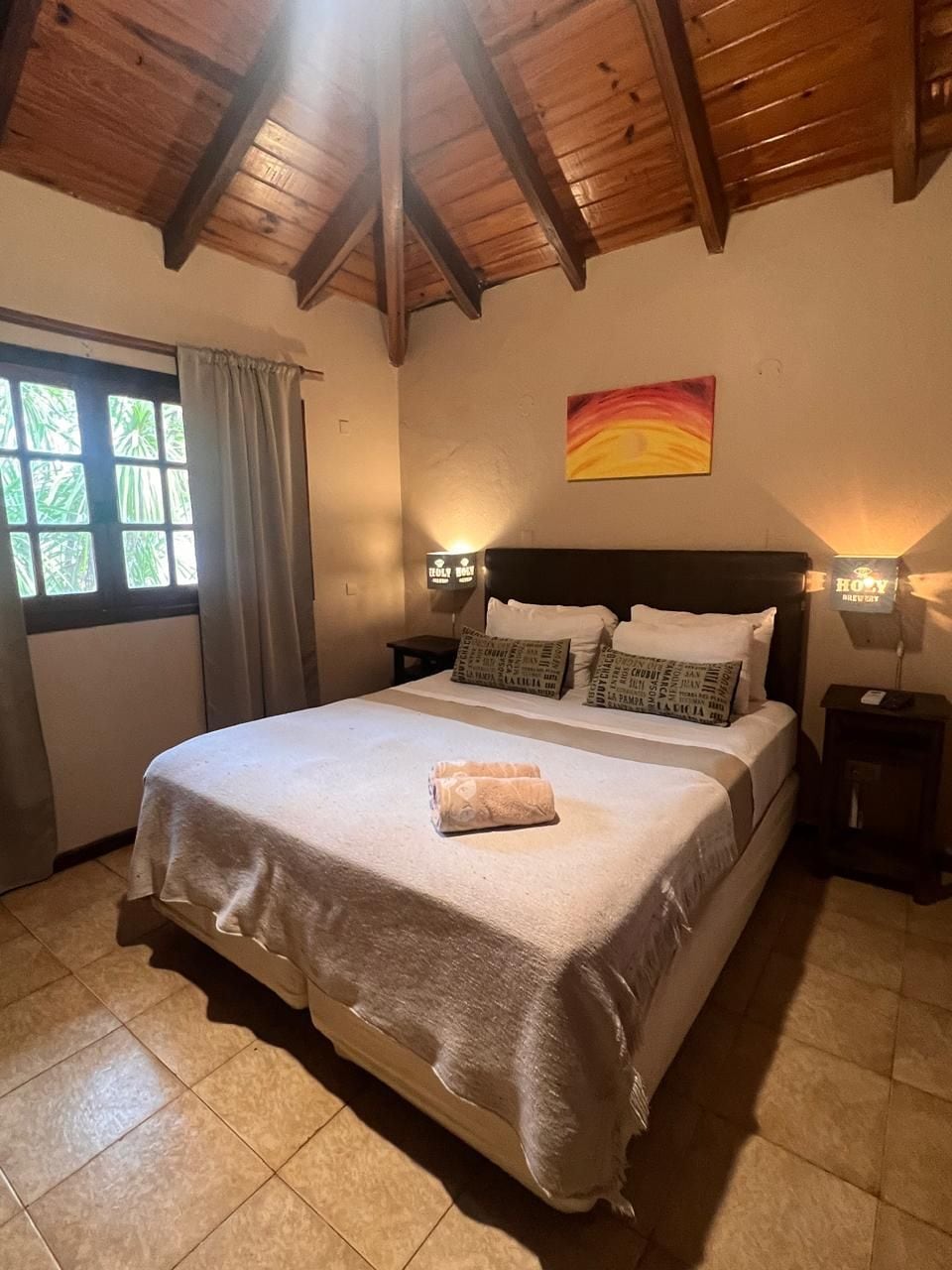 Ein Schlafzimmer mit einem großen Bett, Holzbalkendecke, einem Fenster mit Vorhängen und Wandlampen.