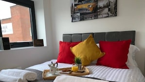 Et hotelværelse med seng, puder, et bakke med morgenmad, et vindue og en væg med et bybillede.