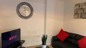En stue med et fladskærms-tv, der viser 'NETFLIX', en sort lædersofa med røde puder, et lille sofabord med en plante og et vægur.