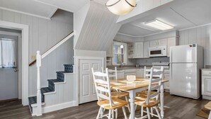 Una cocina con armarios blancos, un refrigerador blanco, una mesa de comedor de madera con sillas y una escalera con peldaños estampados.